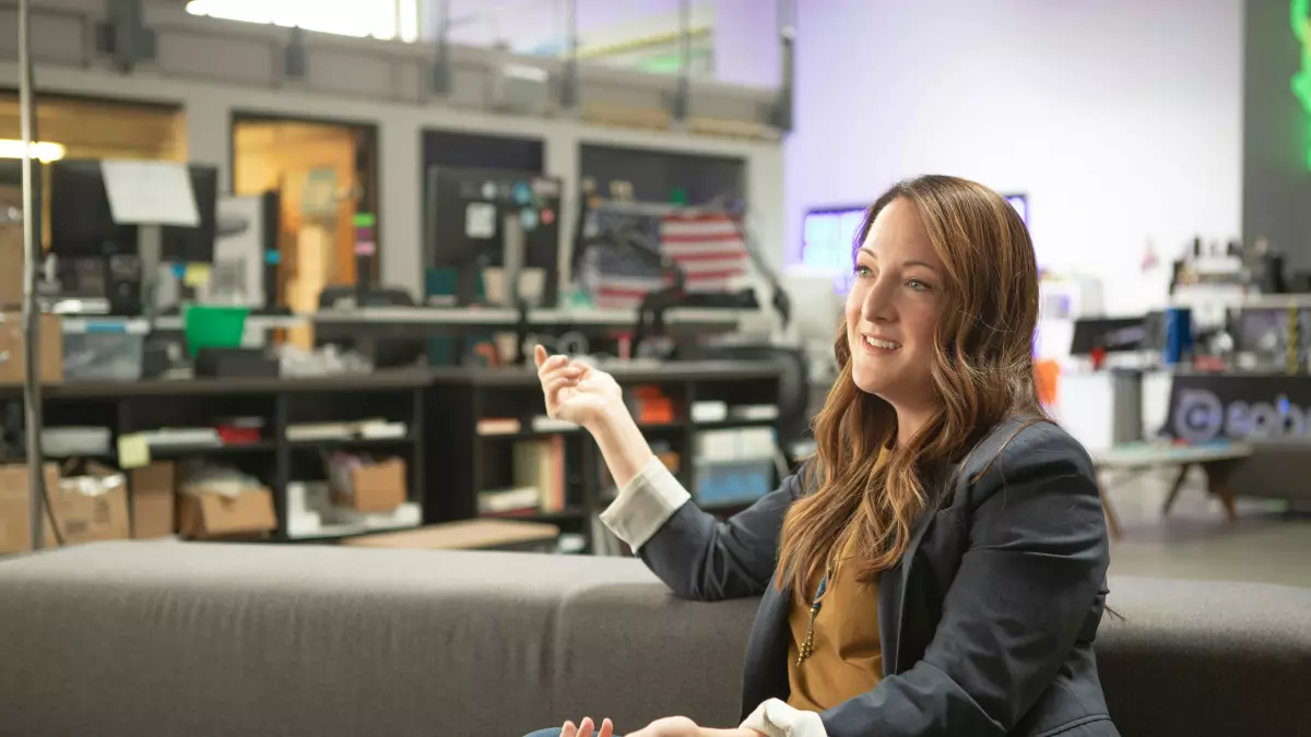 A woman sitting on a couch in an office setting, gesturing with her hands and smiling, possibly talking about a project.