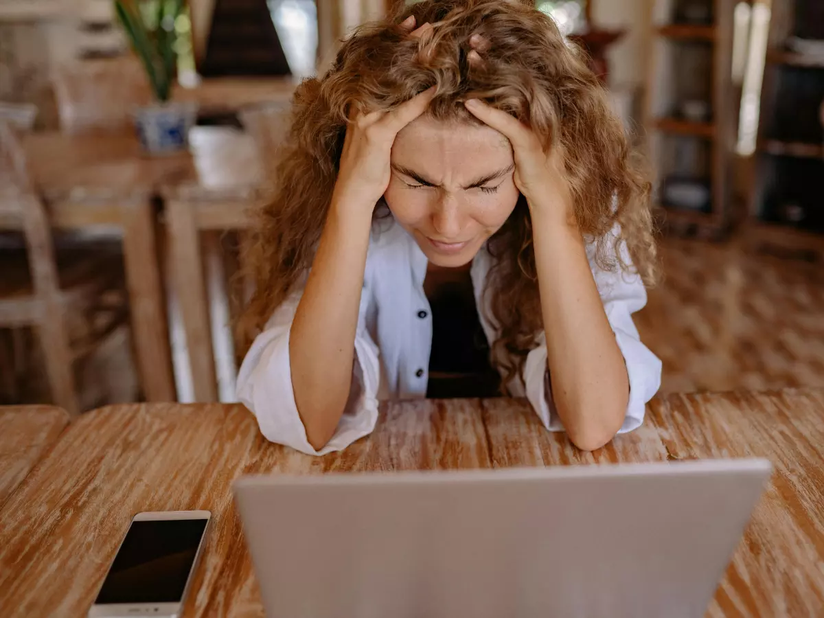 A woman sitting at a table in front of a laptop, with her hands in her hair, looking frustrated.