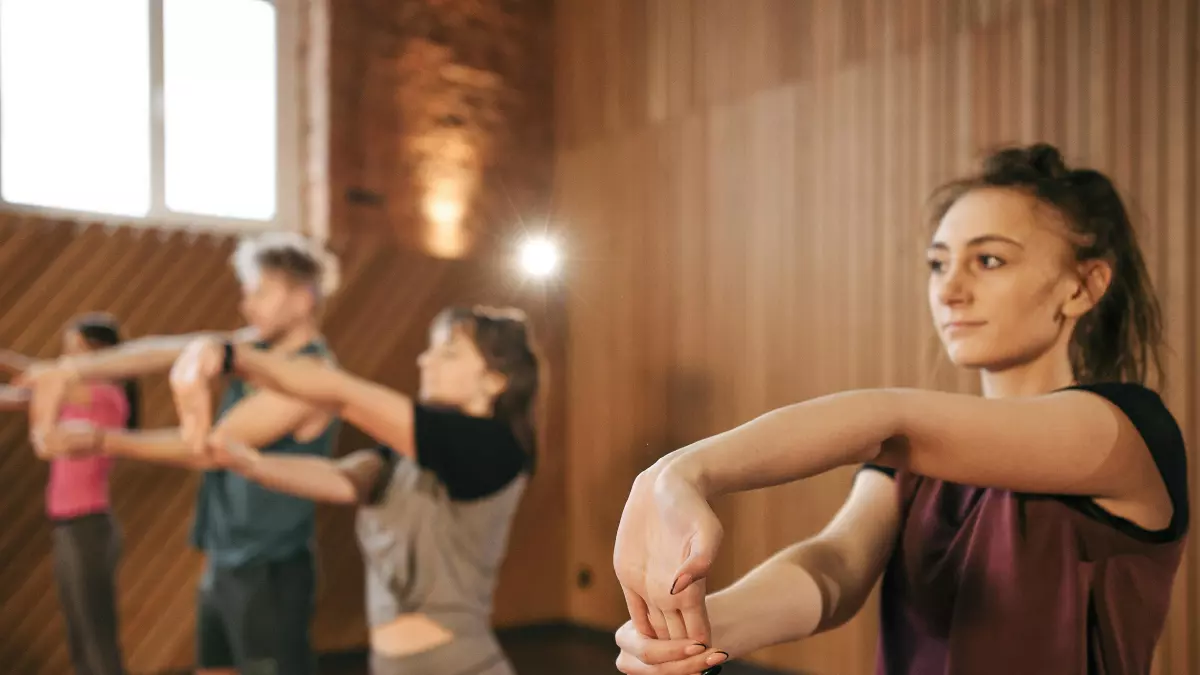 A young woman in a dark red tank top with a ponytail stretches her arms in a dance studio. She is at the forefront, while other dancers are blurred in the background.