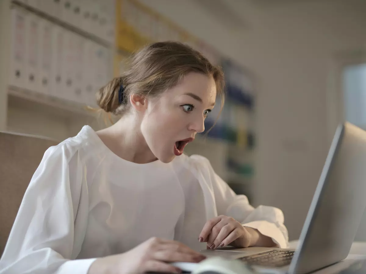 A woman is sitting at a desk and looking at a laptop screen. She has a surprised expression on her face. She is wearing a white shirt and has her hair pulled back.