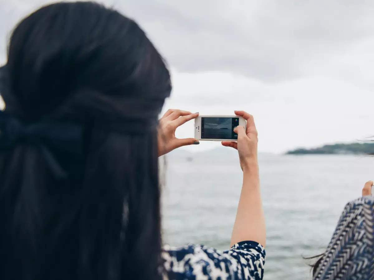 Two women standing on a rocky beach, looking out at the sea.  The woman in the foreground is holding a smartphone in her hands, and she is taking a picture.