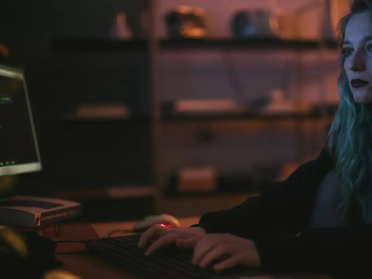 A woman with long blond hair sits in front of a computer, typing on a keyboard. She's wearing a black hoodie, and the room is dimly lit. The screen is displaying a blurry image.