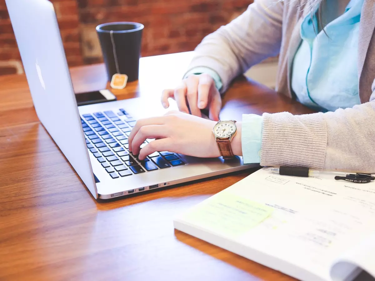 Close-up image of a woman's hands typing on a laptop with a notebook and pen on the desk.