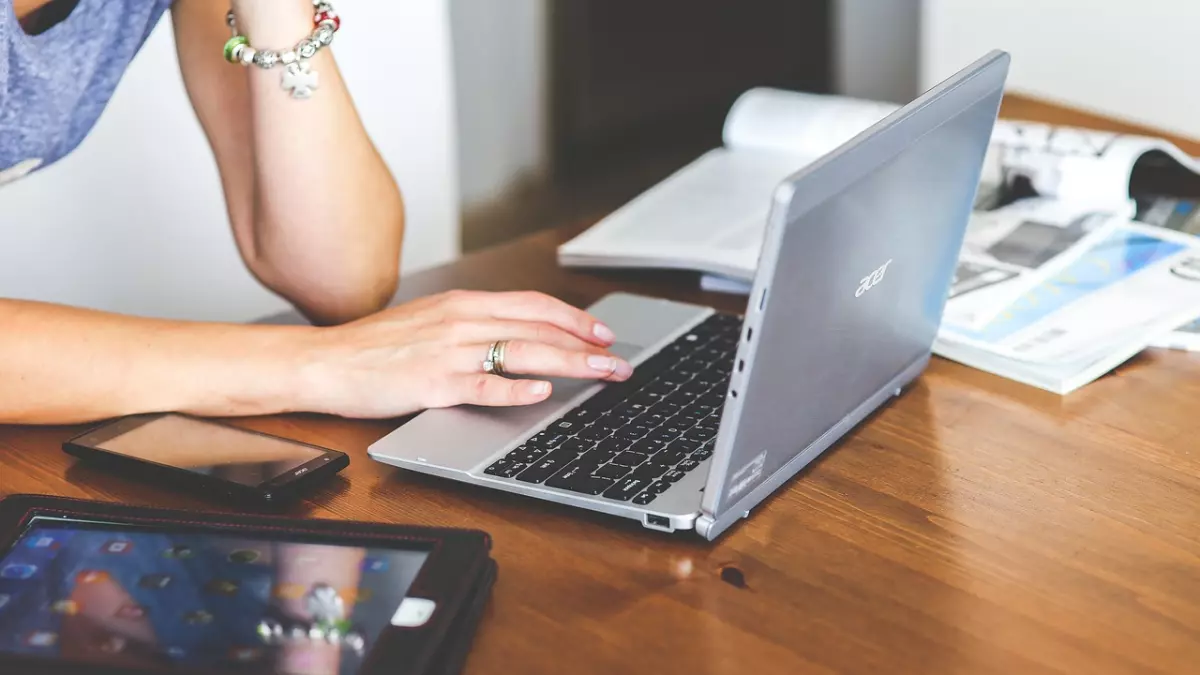 A person sitting at a desk, using a laptop. There are other electronic devices on the desk, including a phone and a tablet.