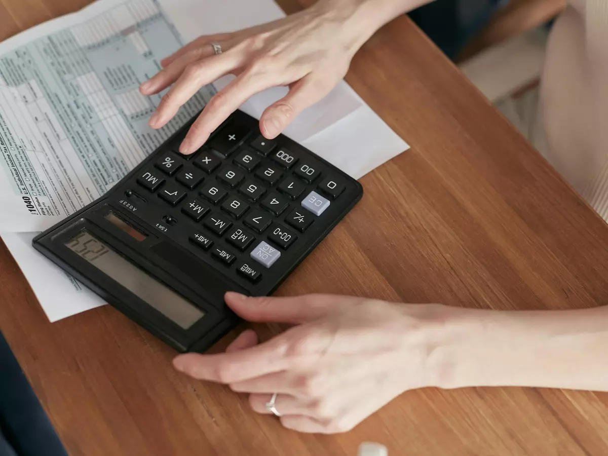 A woman is holding a calculator. She is wearing a white shirt and her hand is on the calculator. There are some papers on the table.