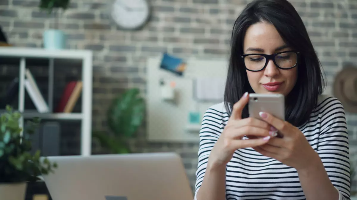 A woman wearing glasses uses her phone while sitting at a desk. 