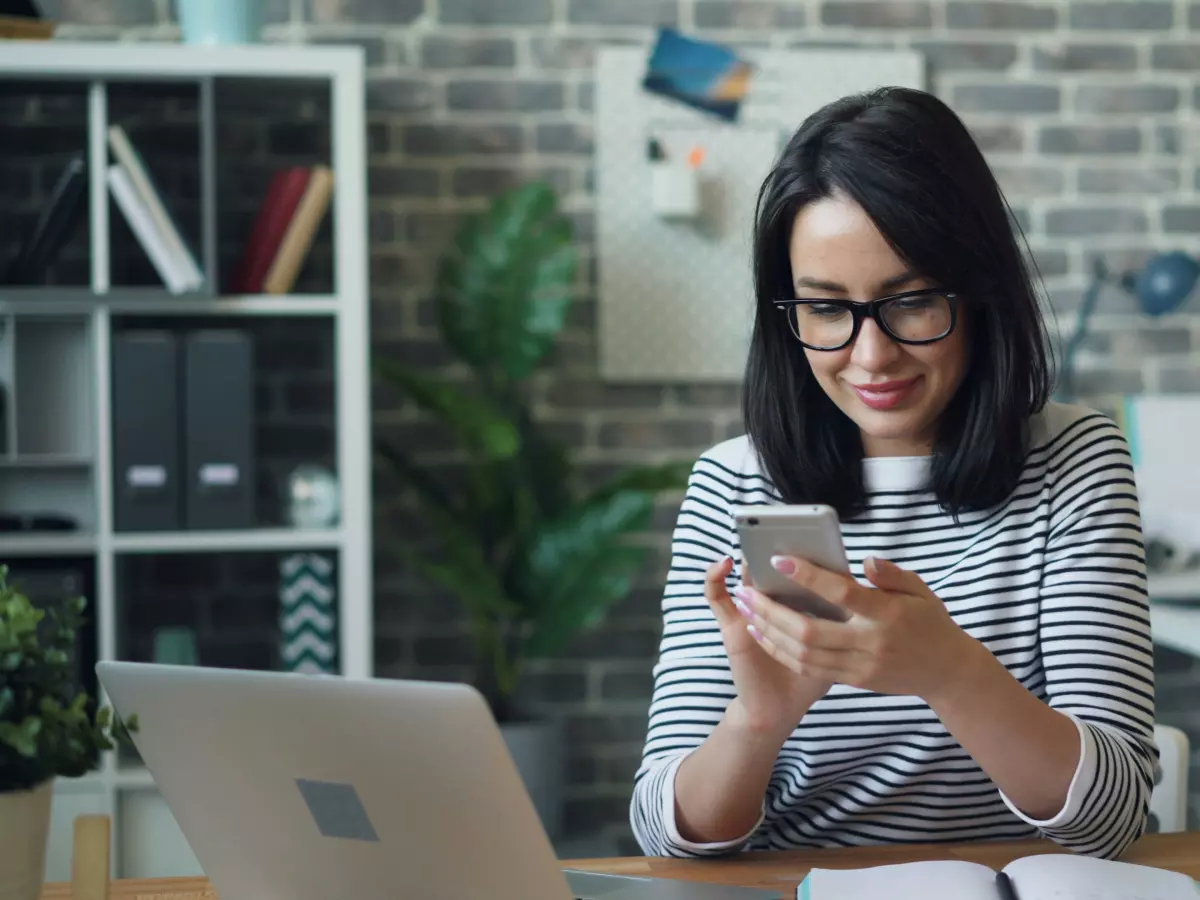 A woman with dark hair and glasses is smiling while looking at her phone, with a laptop and a notepad on the table in front of her.