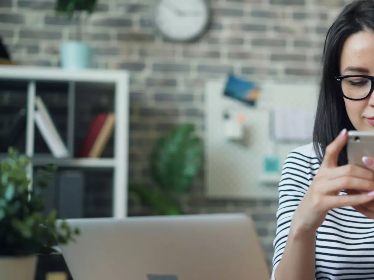 A woman wearing glasses uses her phone while sitting at a desk. 