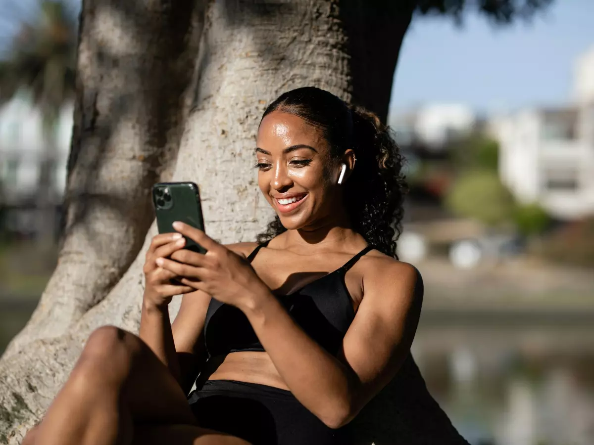 A woman in a black sports bra and shorts is sitting under a tree, smiling and looking at her phone.