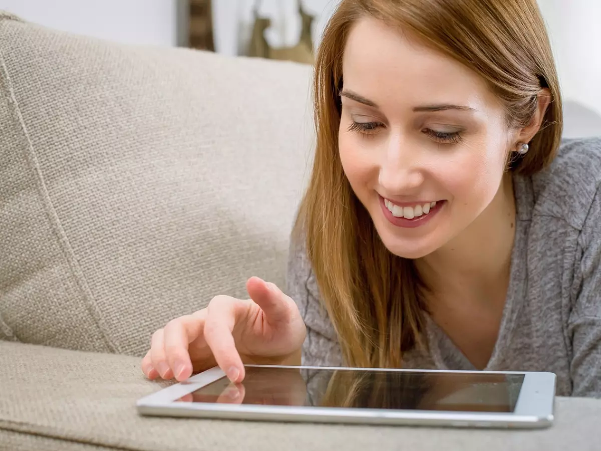 A woman is lying on a couch and using a tablet, she is smiling.