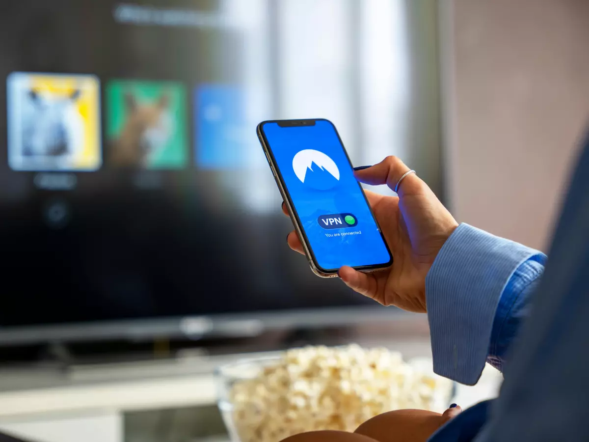 A man is holding a phone with a blue screen and a VPN logo. He is looking at the screen and there is a TV in the background with a blurry image of a video streaming service. The man is sitting in a living room with popcorn in front of him.