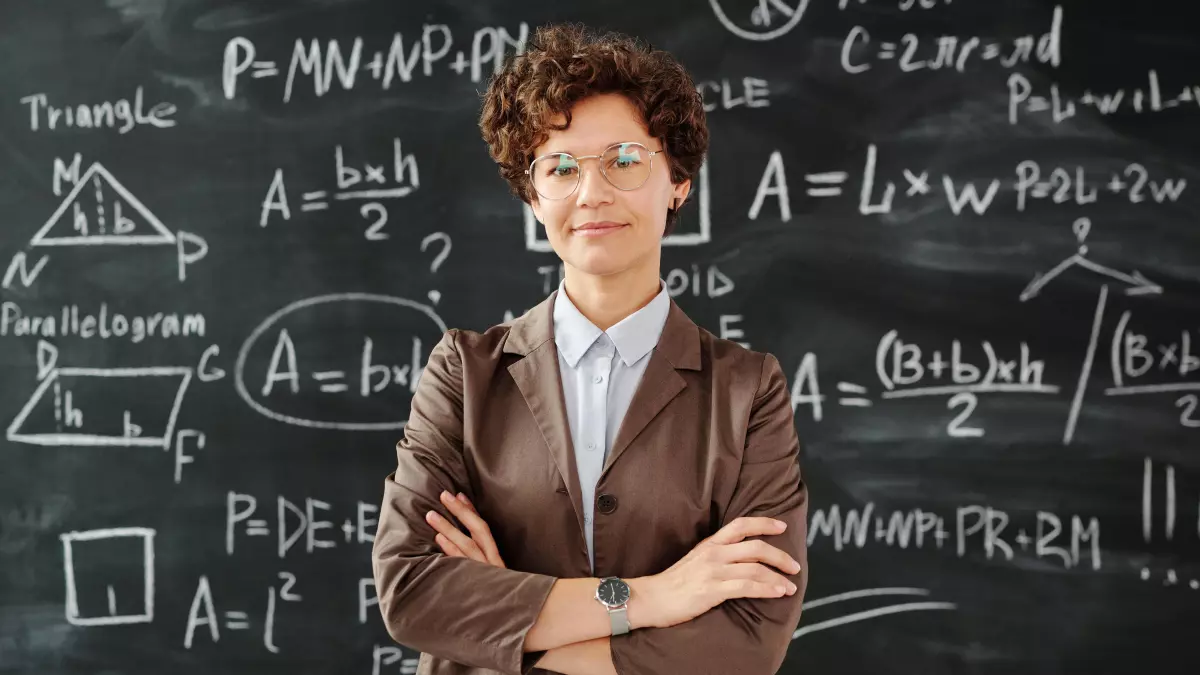 A woman in a brown blazer stands in front of a blackboard covered in handwritten math equations. She has short curly hair, and she is looking directly at the camera. Her arms are crossed in front of her.