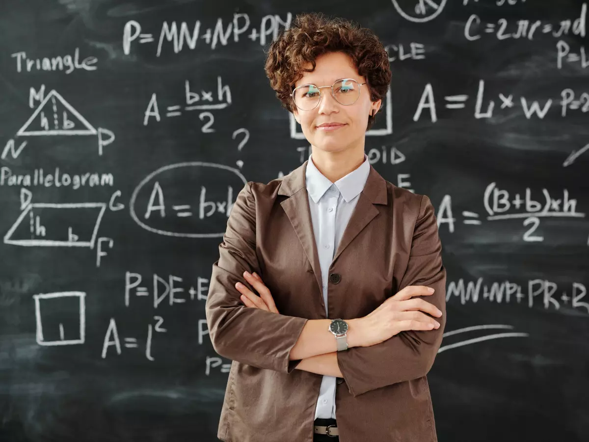 A woman in a brown blazer stands in front of a blackboard covered in handwritten math equations. She has short curly hair, and she is looking directly at the camera. Her arms are crossed in front of her.