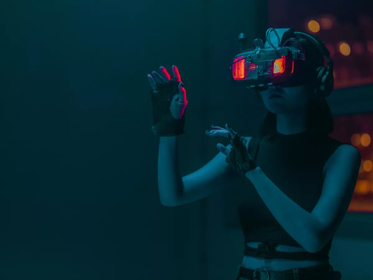 A woman wearing VR goggles, with red LED lights illuminating her face, holds out her hands in a gesture of wonder or surprise.