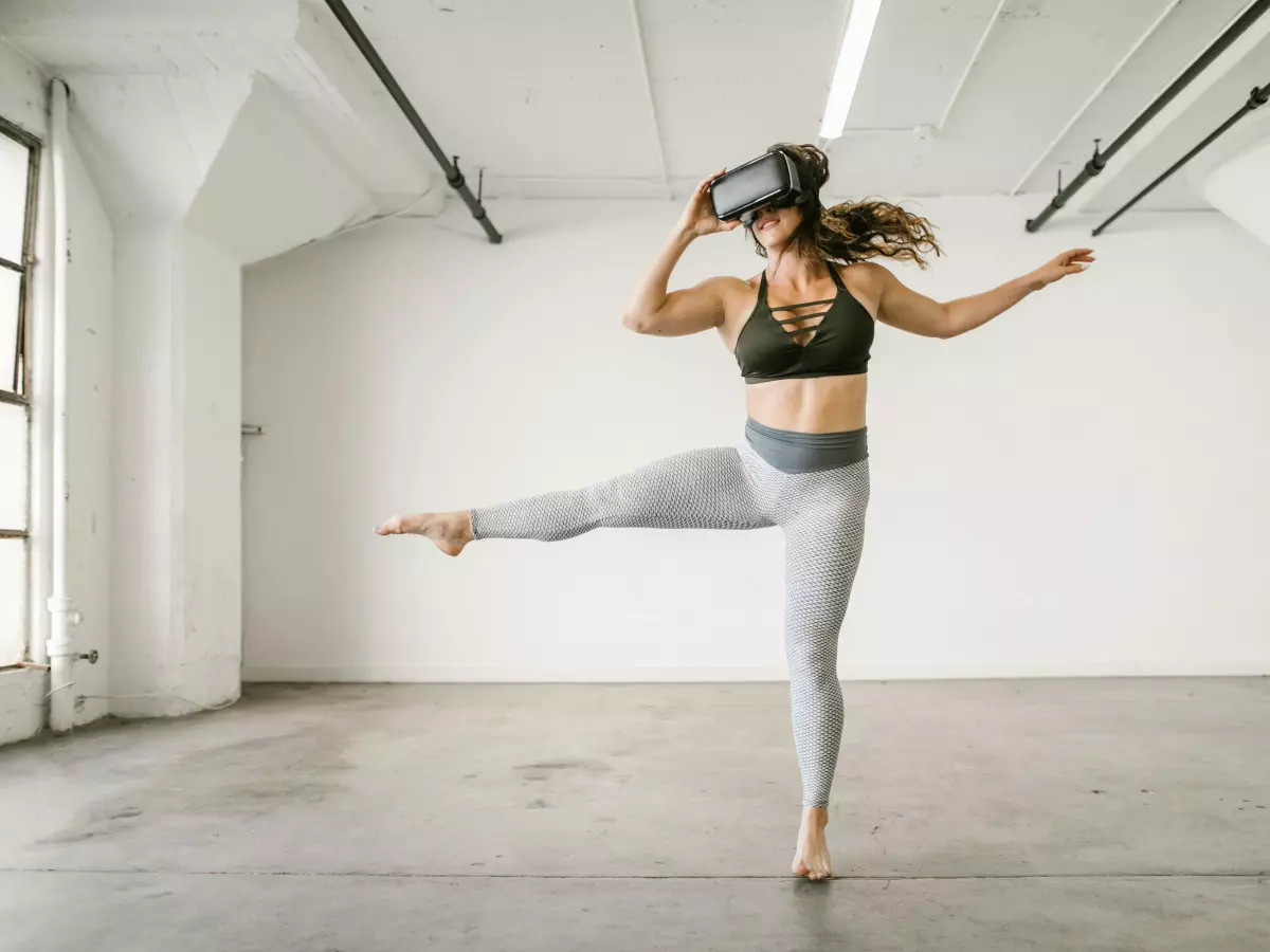 A woman wearing a VR headset is kicking her leg high, a dynamic pose in a dance studio setting.