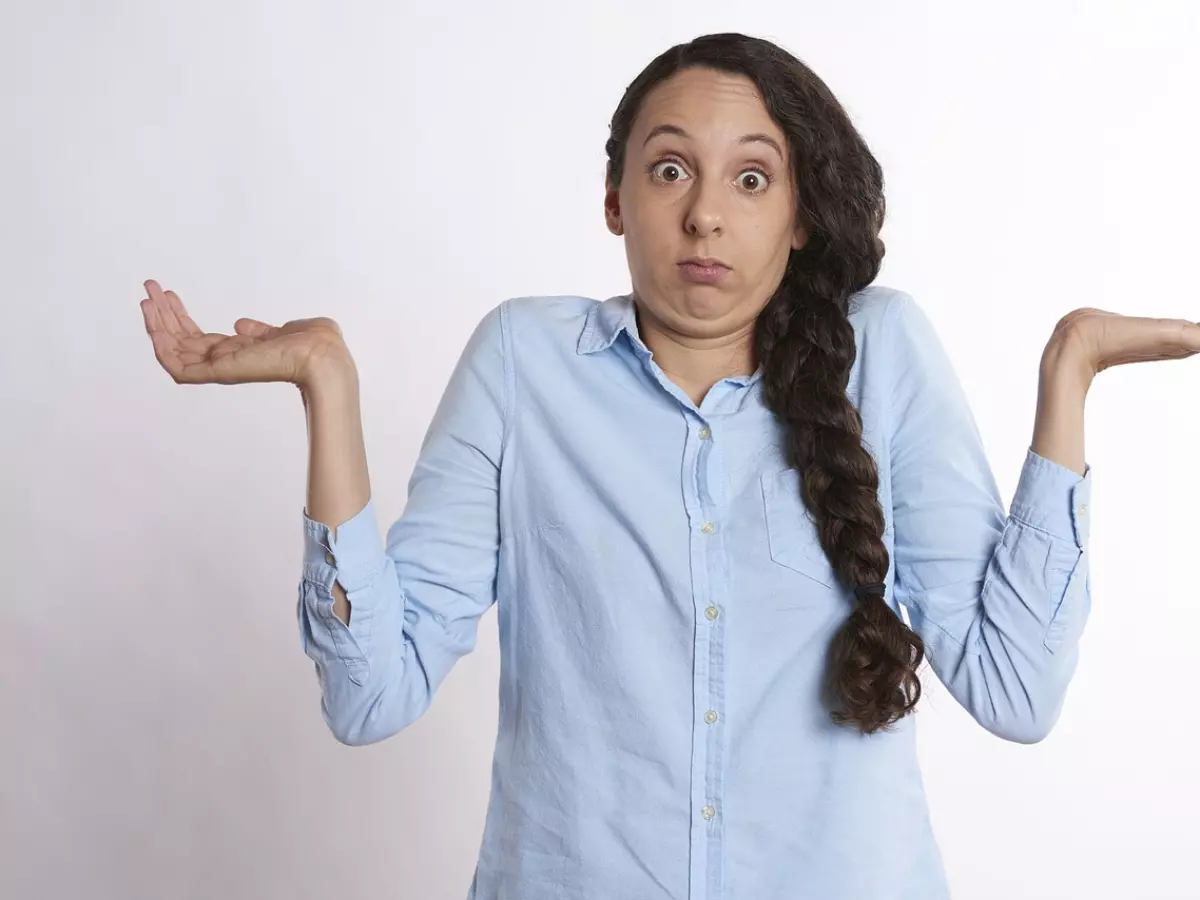 A woman with long brown hair is standing against a white background with her arms out and a confused expression on her face.