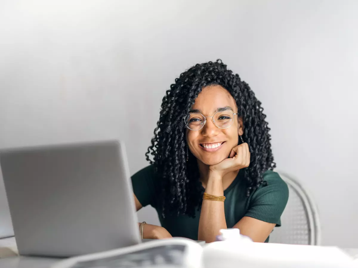 A young woman is smiling while looking at a laptop, she has her hand on her chin, she is sitting with a book next to her.