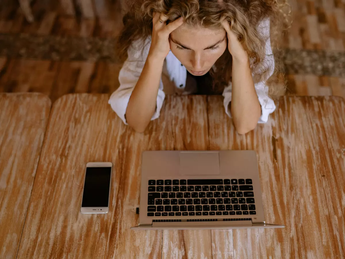 A young woman, wearing a white blouse and sitting on a wooden floor, holds her head in her hands in front of an open laptop with a smartphone next to it.