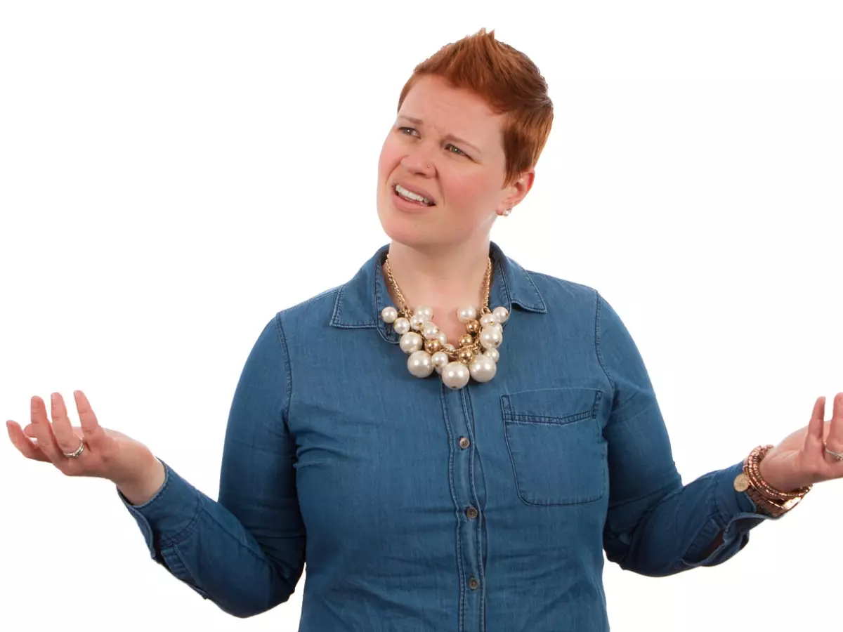 A woman with red hair and a blue shirt is looking up with a confused expression and her hands are raised in the air. She is wearing a pearl necklace and multiple bracelets.