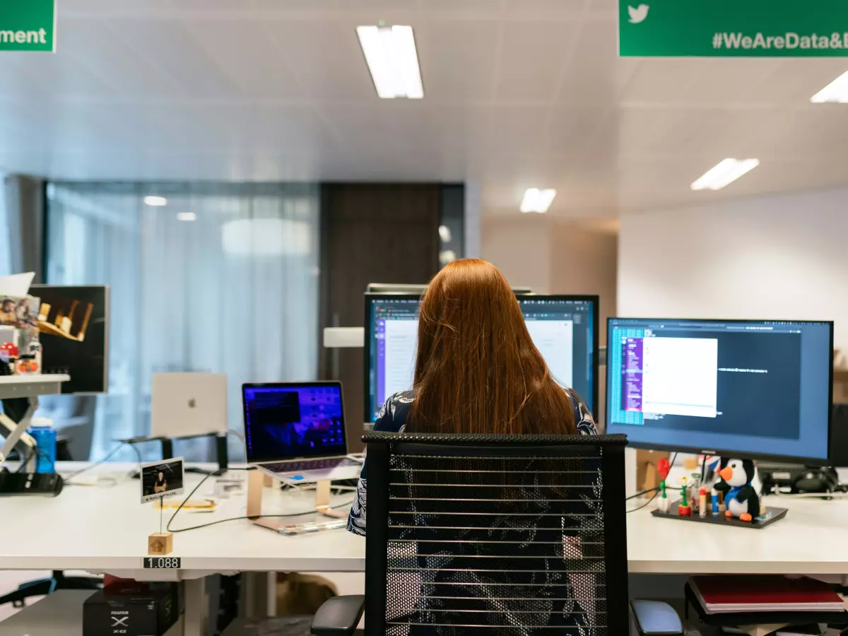 A woman sits at a desk with multiple monitors, typing on her laptop keyboard.