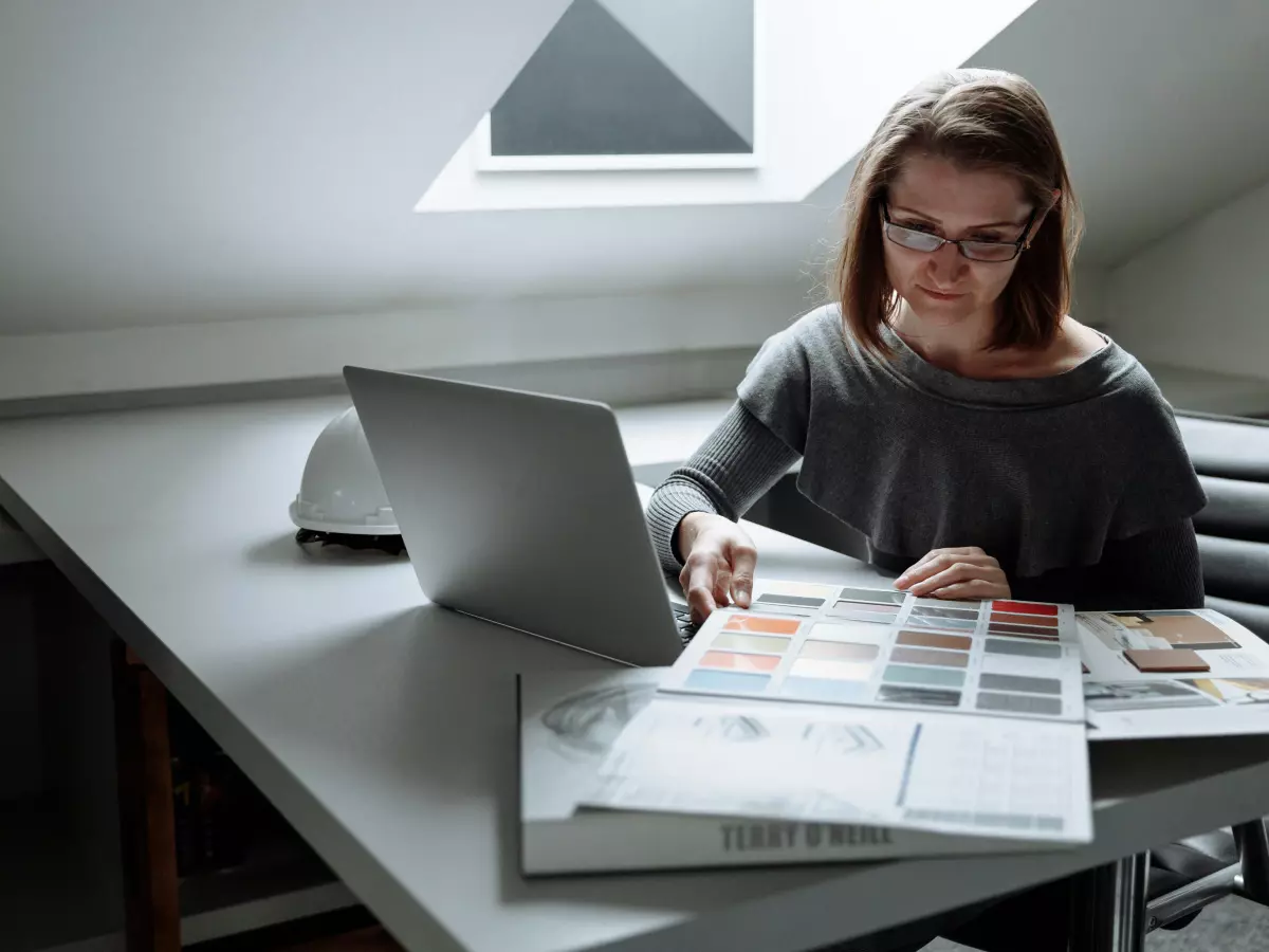 A woman sitting at a desk with a laptop and a spread of design work, she is looking down at the paper.