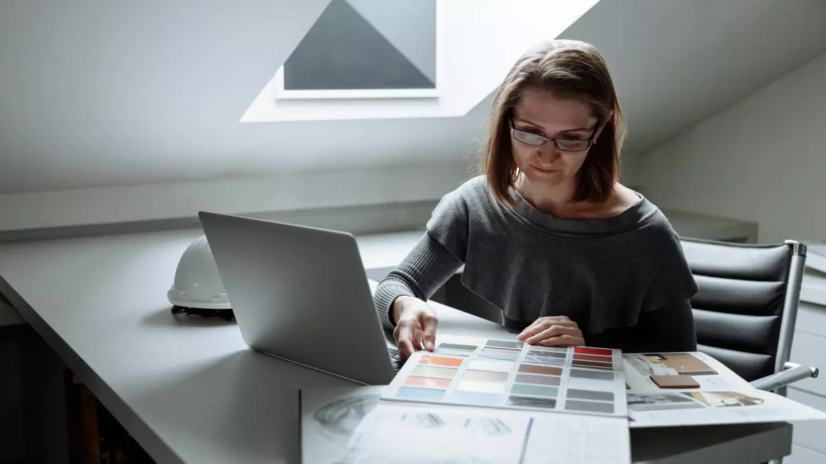 A woman sitting at a desk with a laptop and a spread of design work, she is looking down at the paper.