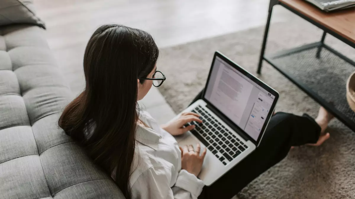 A woman sits on the floor, back to the camera, using a laptop. 