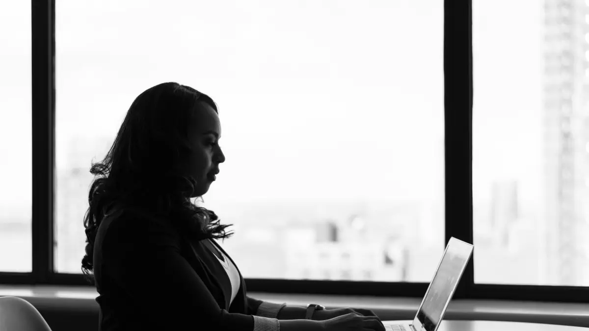 A woman sits at a table in an office, working on a laptop. She is wearing a blazer and her hair is pulled back. She is looking at the screen and her hands are typing on the keyboard. There is a large window behind her, and the light from outside makes the image slightly darker. The focus is on her and the laptop.