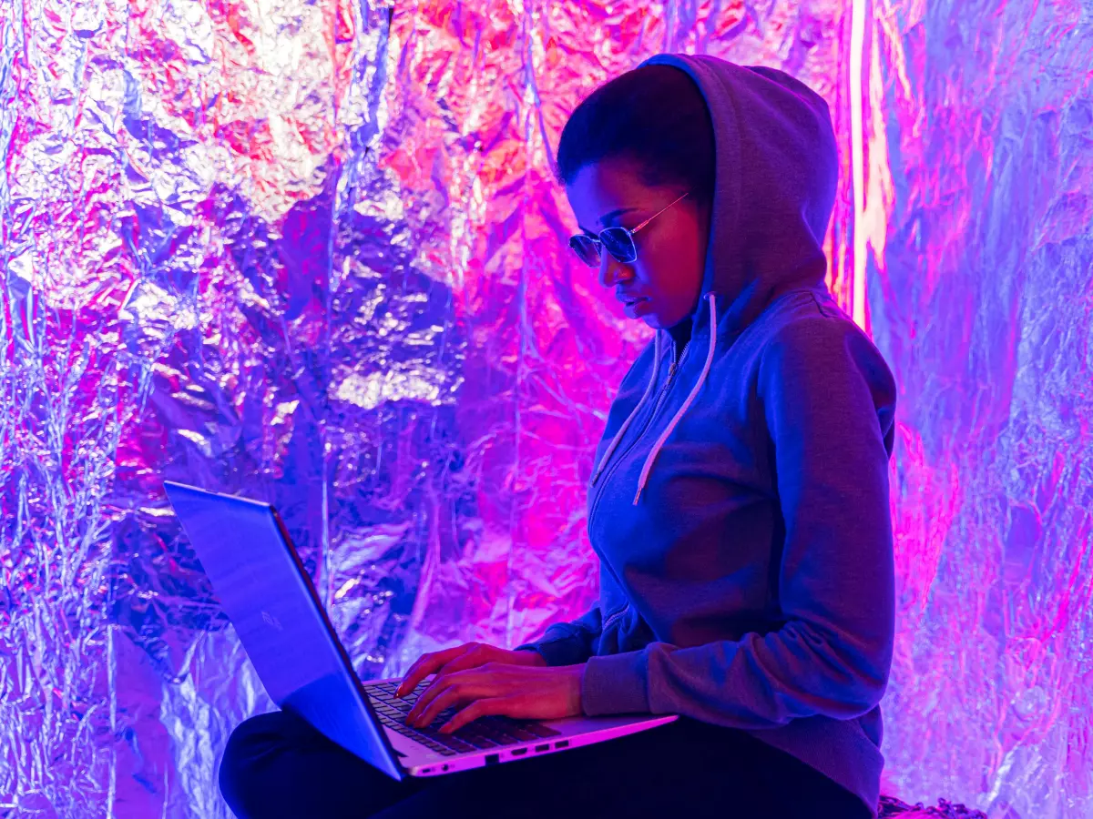 A person wearing a black hoodie, mask, and glasses, in a dimly lit room, possibly engaging in online activities, evoking the stereotypical hacker image.