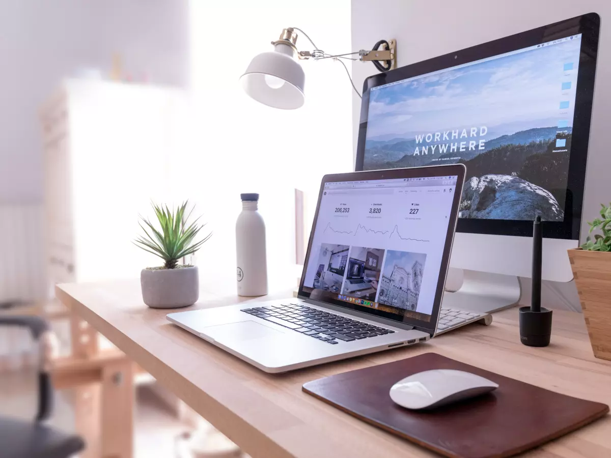 A laptop and a desktop computer on a wooden table, both displaying a website. The laptop is in focus, and the desktop is in the background.