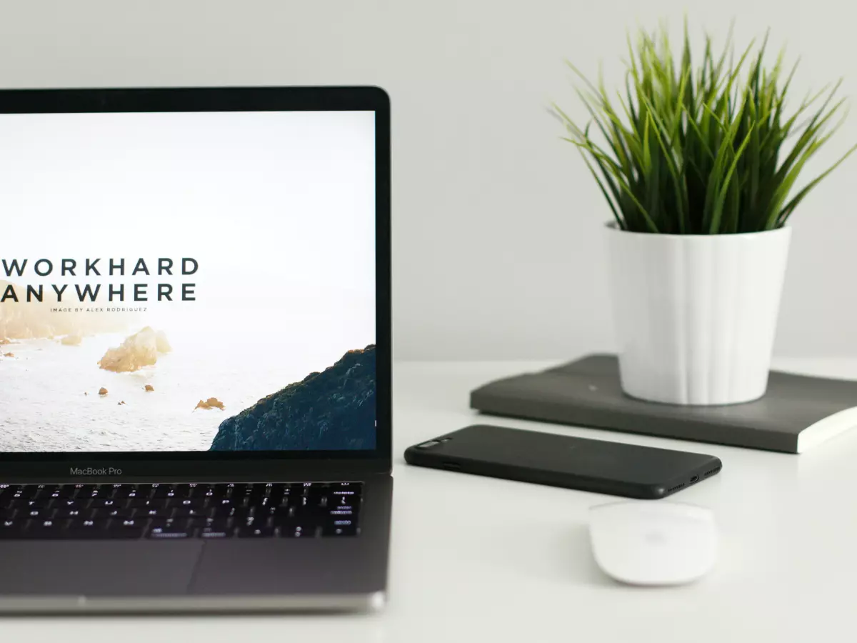 A laptop, phone, and mouse on a white desk. There is a potted plant behind the laptop.