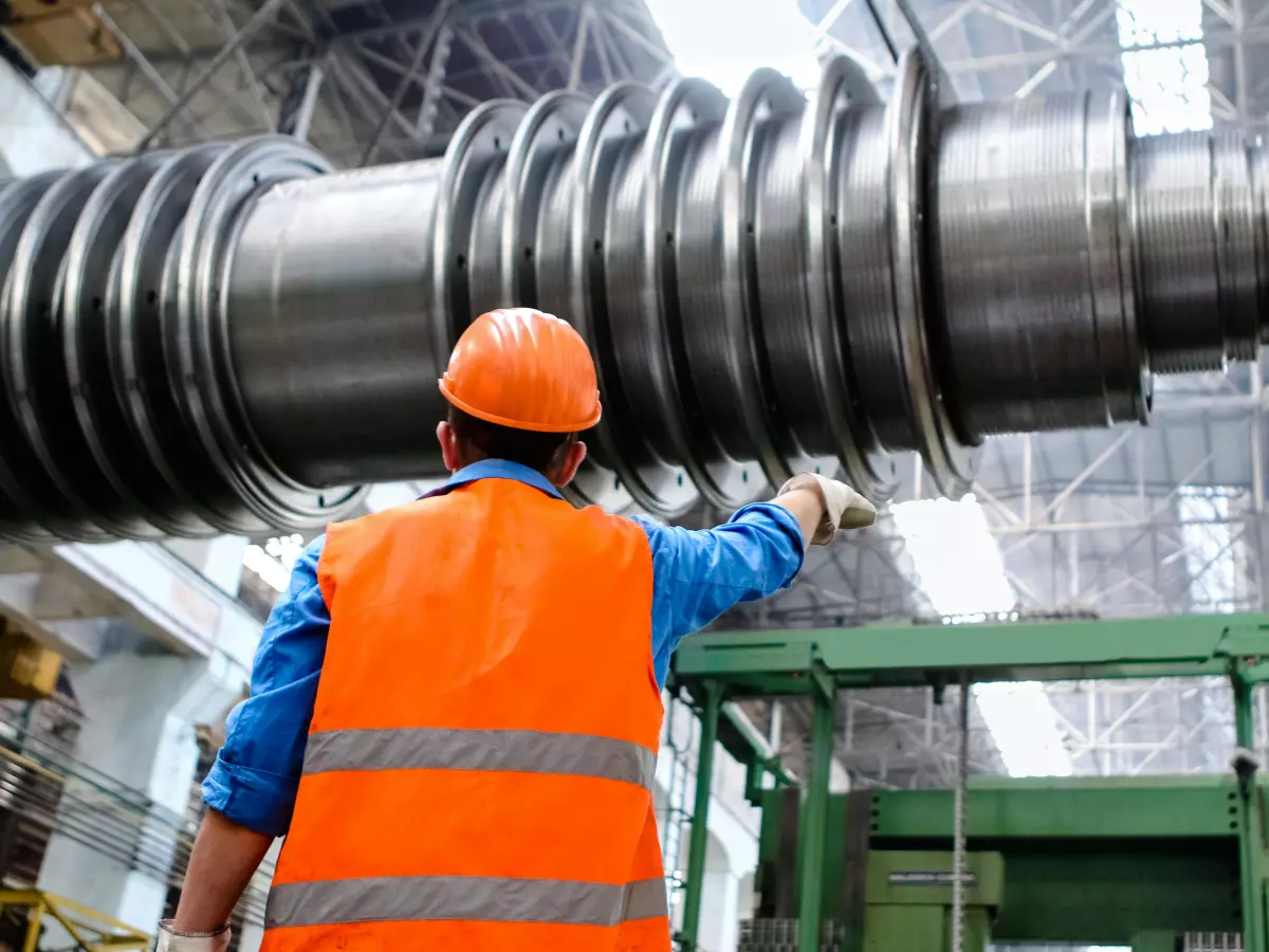 A construction worker with a hard hat and safety vest points at something in the distance while standing on a platform in a factory setting. The image conveys a sense of scale and complexity.