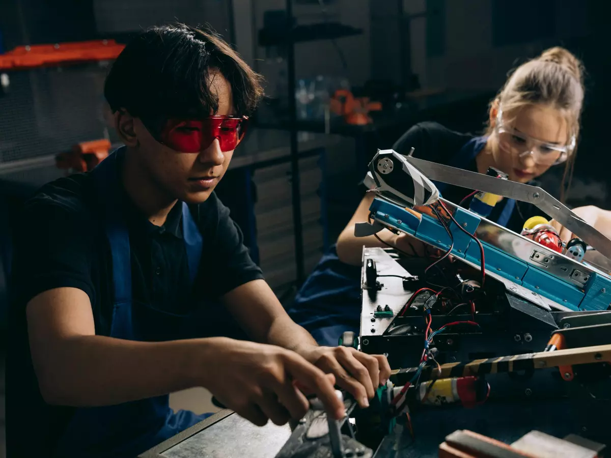 Two young people work together on a robot in a garage, their faces are lit by red and blue light.