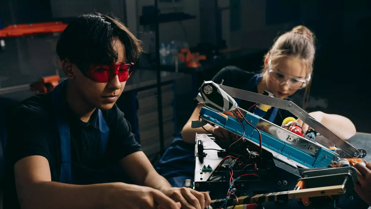 Two young people work together on a robot in a garage, their faces are lit by red and blue light.