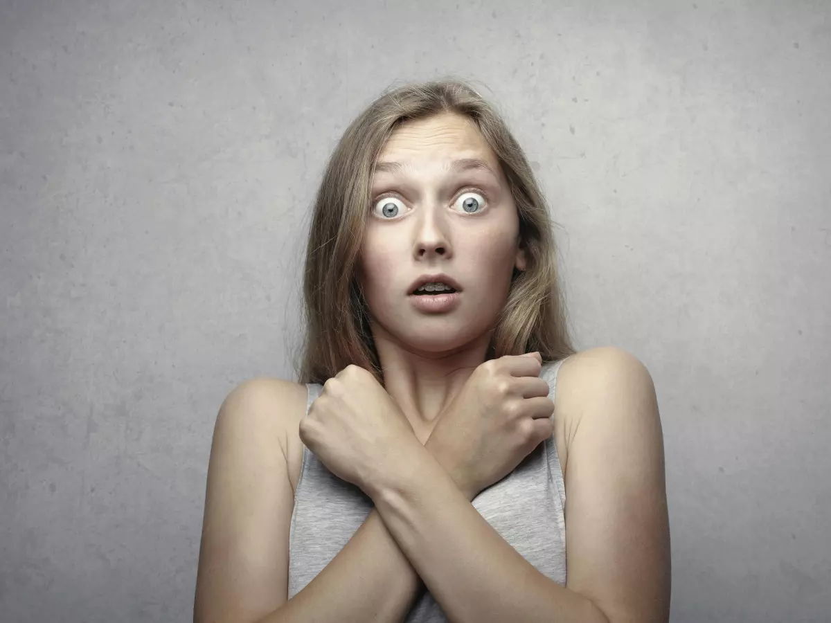 A woman with wide eyes and a scared expression. Her hair is light brown, and she is wearing a white tank top, her arms are crossed. The background is plain and unassuming.