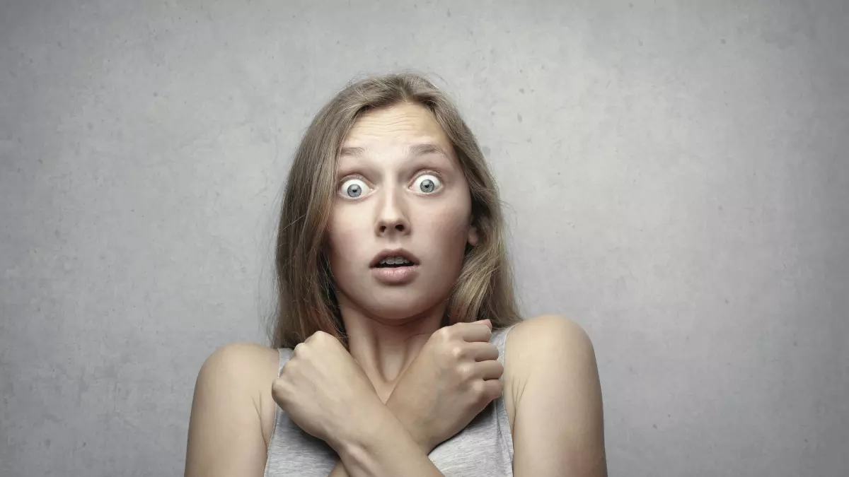 A woman with wide eyes and a scared expression. Her hair is light brown, and she is wearing a white tank top, her arms are crossed. The background is plain and unassuming.